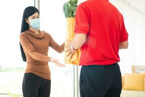 Asian woman wearing a face mask and pickup shopping bag of Fresh food, vegetables, and fruits from a delivery man from a supermarket. Concept of express grocery service and new  lifestyle photo