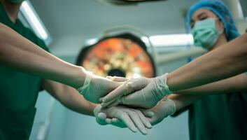 asiático cirujano y enfermero con médico máscara en pie y unirse mano en operación teatro a un hospital. equipo de profesional cirujanos cuidado de la salud, emergencia médico Servicio concepto foto