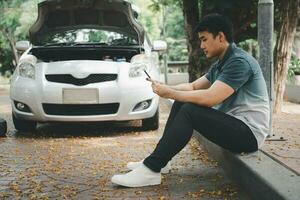 Asian man using smartphone for assistance after a car breakdown on street. Concept of vehicle engine problem or accident and emergency help from Professional mechanic photo