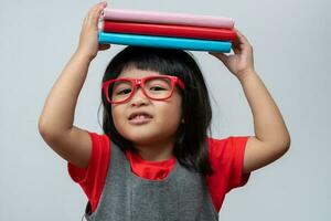 Funny and Happy Asian little preschool girl wearing red glasses holding a green book on the head, on white isolated background. Concept of school kid and education in elementary and preschool photo