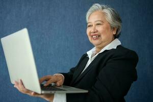 Portrait of intelligent senior businesswoman CEO manager holding laptop creating a presentation on isolated blue background. Old Woman is happy and smile after getting financial statements reports photo