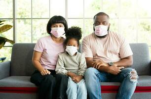 Mixed Race family sitting on the sofa and wearing medical face masks for against coronavirus world pandemic and stay quarantined together at home social distancing new normal lifestyle photo