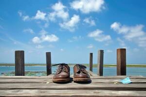 un par de marrón cuero zapatos, con cordones, ese tener estado izquierda en un muelle cerca un cuerpo de agua. el Zapatos parecer a ser viajando desde el mar a el cielo, simbolizando personal crecimiento y cambiar. foto