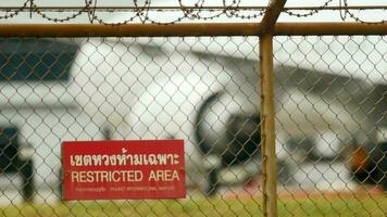 PHUKET, THAILAND NOVEMBER 29, 2018 - A metallic fence surrounds the airport area. Airplane is taxiing to the runway in soft focus. There is a sign Restricted area on the fence video