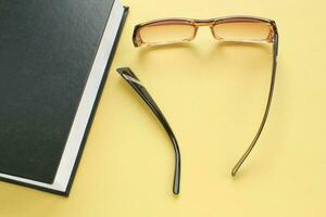 Old broken eyeglasses with damaged legs and books on yellow background. Poor eyesight. Repair concept. Idea of health. Failure optic eyewear. Breakage of vision correction glasses. Close up, flat lay photo