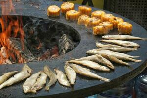 Grilled fish and corn on a bowl-shaped flat grill with fire hole in the center. Outdoor seafood barbecue cooking. Picnic on backyard on open air. Street food concept. Preparing fish over open campfire photo