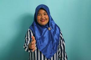 A confident middle-aged Asian woman, wearing a blue hijab and a striped shirt, invites you to shake hands while standing against a blue background photo