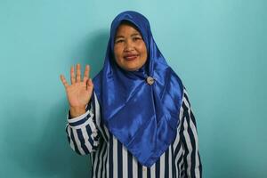 A friendly middle-aged Asian woman in a blue hijab and striped shirt is smiling, saying hi, and waving her hand to greet a person, making a hello gesture while standing against a blue background. photo