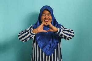 A smiling middle-aged Asian woman, in a blue hijab and striped shirt, makes a heart gesture with her hands, radiating happiness and care, isolated on blue background photo