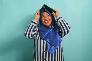 The happy Asian middle-aged woman, donning a blue hijab and a striped shirt, holds a book above her head like a makeshift roof while standing against a blue background. photo