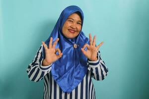 A smiling middle-aged Asian woman in a blue hijab and striped shirt is standing against a blue background, confidently displaying the OK gesture photo