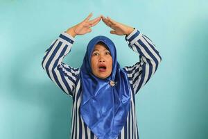 A surprised middle-aged Asian woman in a blue hijab and striped shirt is making a home or roof gesture by holding her hands above her head. She is isolated over a blue background photo