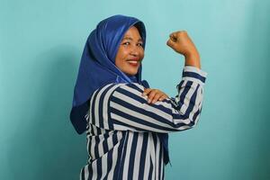 An excited middle-aged Asian woman in a blue hijab and striped shirt shows a strong gesture by lifting her arms and flexing her muscles, smiling proudly at the camera, isolated on blue background. photo