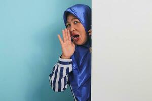 A middle-aged Asian woman in a hijab, amazed, hides behind a white wall with a blank space, peeking out and shouting next to a white advertising board for text design or advertisement. photo