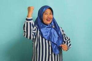 An enthusiastic middle-aged Asian woman, in a blue hijab and a striped shirt, is saying YES with a happy expression, celebrating victory with a fist pump gesture while standing over a blue background photo