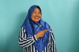 An enthusiastic middle-aged Asian woman, wearing a blue hijab and striped shirt, points to the right, emphasizing the copyspace. Isolated over blue background. photo