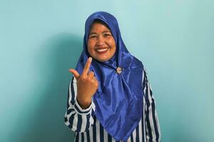 A middle-aged Asian woman, in a blue hijab and striped shirt, confidently makes an inviting or beckoning gesture with her hand, signaling someone to come. Isolated on a blue background photo