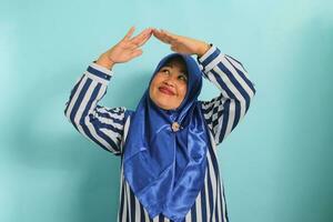 An excited middle-aged Asian woman in a blue hijab and striped shirt is making a home or roof gesture by holding her hands above her head while standing against a blue background photo