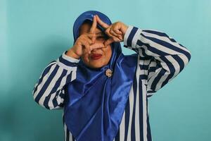 A middle-aged Asian woman in a blue hijab and striped shirt forms a triangle with her fingers while standing against a blue background photo