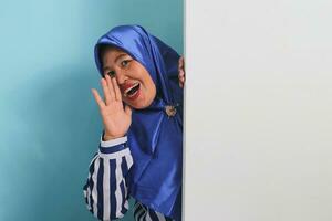 A middle-aged Asian woman in a hijab, amazed, hides behind a white wall with a blank space, peeking out and shouting next to a white advertising board for text design or advertisement. photo