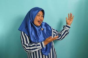 An excited middle-aged Asian woman in a blue hijab and striped shirt shows copy space aside, recommending a great offer or discount with her open palms. She is isolated on a blue background photo