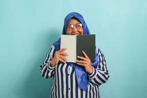 Excited Asian middle-aged woman, wearing a blue hijab, eye glasses and a striped shirt, hides her face behind an open book while looking away, standing against a blue background. photo