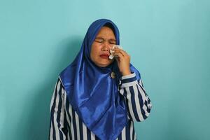 An upset middle-aged Asian woman, wearing a blue hijab and a striped shirt, is crying and wiping tears with a tissue, expressing frustration and distress while standing against a blue background photo
