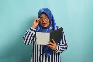 Amazed Asian middle-aged woman, wearing a blue hijab, eyeglasses, and a striped shirt, holds an open book with a surprised expression while standing against a blue background. photo