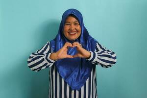 A smiling middle-aged Asian woman, in a blue hijab and striped shirt, makes a heart gesture with her hands, radiating happiness and care, isolated on blue background photo