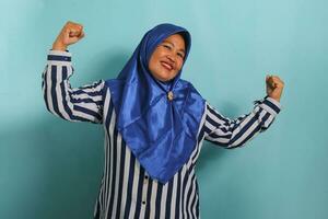 An excited middle-aged Asian woman in a blue hijab and striped shirt shows a strong gesture by lifting her arms and flexing her muscles, smiling proudly at the camera, isolated on blue background. photo