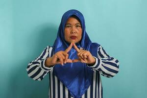 A middle-aged Asian woman in a blue hijab and striped shirt forms a triangle with her fingers while standing against a blue background photo