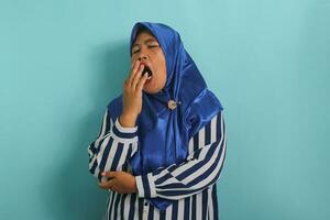 Sleepy Asian woman, middle-aged, wearing blue hijab and striped shirt, yawns and covers mouth, looking bored and tired, standing against blue background. Morning fatigue. photo