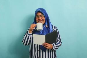 Excited Asian middle-aged woman, wearing a blue hijab, eyeglasses, and a striped shirt, holds an open book and a mug while standing against a blue background. photo