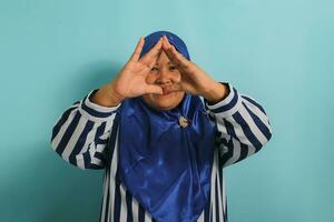 A middle-aged Asian woman in a blue hijab and striped shirt forms a triangle with her fingers while standing against a blue background photo