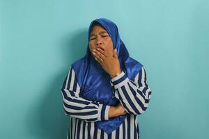 Sleepy Asian woman, middle-aged, wearing blue hijab and striped shirt, yawns and covers mouth, looking bored and tired, standing against blue background. Morning fatigue. photo