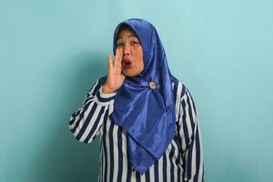 An excited middle-aged Asian woman in a blue hijab and striped shirt shouts and announce something while standing against a blue background. photo