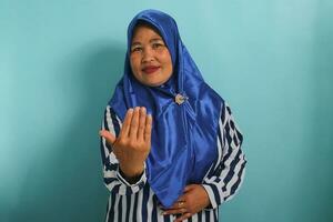A middle-aged Asian woman, in a blue hijab and striped shirt, confidently makes an inviting or beckoning gesture with her hand, signaling someone to come. Isolated on a blue background photo