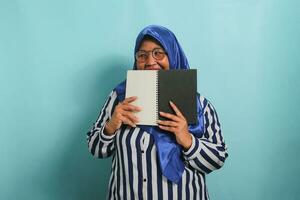 Excited Asian middle-aged woman, wearing a blue hijab, eye glasses and a striped shirt, hides her face behind an open book while looking away, standing against a blue background. photo