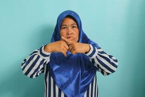 A serious middle-aged Asian woman in a blue hijab and striped shirt makes a cross fingers gesture, indicating to stop talking or stay mute, while standing against a blue background. photo