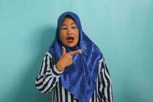 A surprised middle-aged Asian woman in a blue hijab and striped shirt points her fingers to the right while standing against a blue background. photo