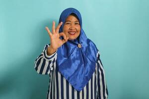 A smiling middle-aged Asian woman in a blue hijab and striped shirt is standing against a blue background, confidently displaying the OK gesture photo