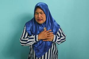 A suffering middle-aged Asian woman, in a blue hijab and striped shirt, clutches her chest in a heart attack gesture, expressing illness and poor health, isolated on a blue background photo