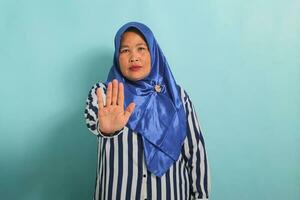 A serious middle-aged Asian woman in a blue hijab and striped shirt shows a stop or refusal gesture while standing against a blue background photo