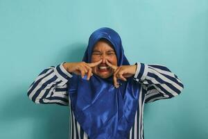 A middle-aged Asian woman, in a blue hijab, holds her breath and pinches her nose with her fingers, reacting to a stinky and disgusting, intolerable bad smell, while standing against a blue background photo