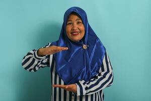 un de edad mediana asiático mujer en un azul hijab y a rayas camisa gestos con su manos, demostración un grande y grande Talla firmar y un medida símbolo. ella es aislado en un azul antecedentes foto