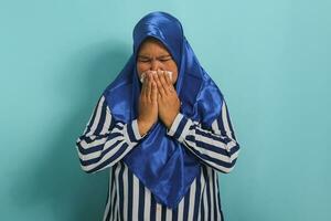un enfermo de edad mediana asiático mujer, vistiendo un azul hijab y un a rayas camisa, es tos y estornudos dentro un pañuelo de papel papel mientras en pie en contra un azul antecedentes foto