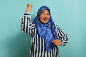 An enthusiastic middle-aged Asian woman, in a blue hijab and a striped shirt, is saying YES with a happy expression, celebrating victory with a fist pump gesture while standing over a blue background photo