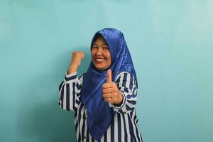 un emocionado de edad mediana asiático mujer en un azul hijab y a rayas camisa muestra un Pulgares hacia arriba y un bueno firmar, simbolizando éxito y expresando aprobación, mientras en pie en contra un azul antecedentes. foto