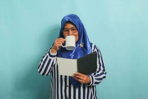 Calm Asian middle-aged woman, wearing a blue hijab, eyeglasses, and a striped shirt, holds an open book and a mug while standing against a blue background. photo