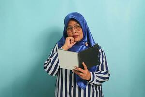 Thoughtful Asian middle-aged woman, wearing a blue hijab and a striped shirt, holds an open book, deep in thought, while standing against a blue background. photo